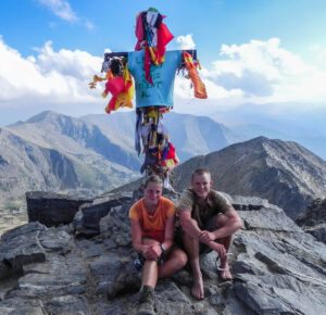 Two people sitting on the summit of the peak infron of the summit cross with some cloth on it